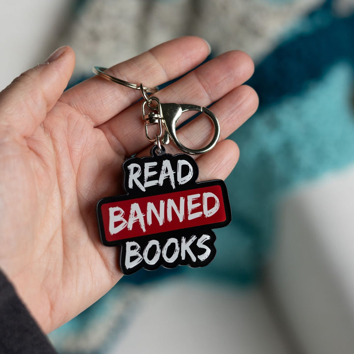 Acrylic keychain that says ‘Read banned books’ in white lettering outlined in black. The word banned is red held in a palm.