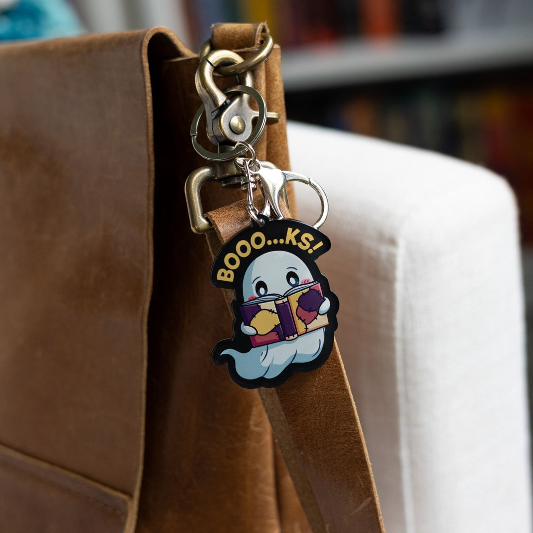  Acrylic keychain of a ghost reading a book with the words ‘Booo…ks!’ over it in yellow clipped to a leather purse.
