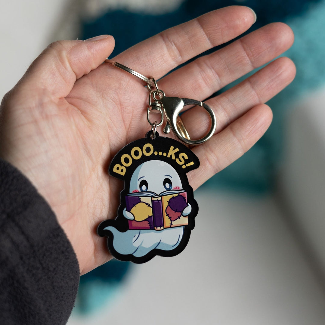 Acrylic keychain of a ghost reading a book with the words ‘Booo…ks!’ over it in yellow held in a palm.