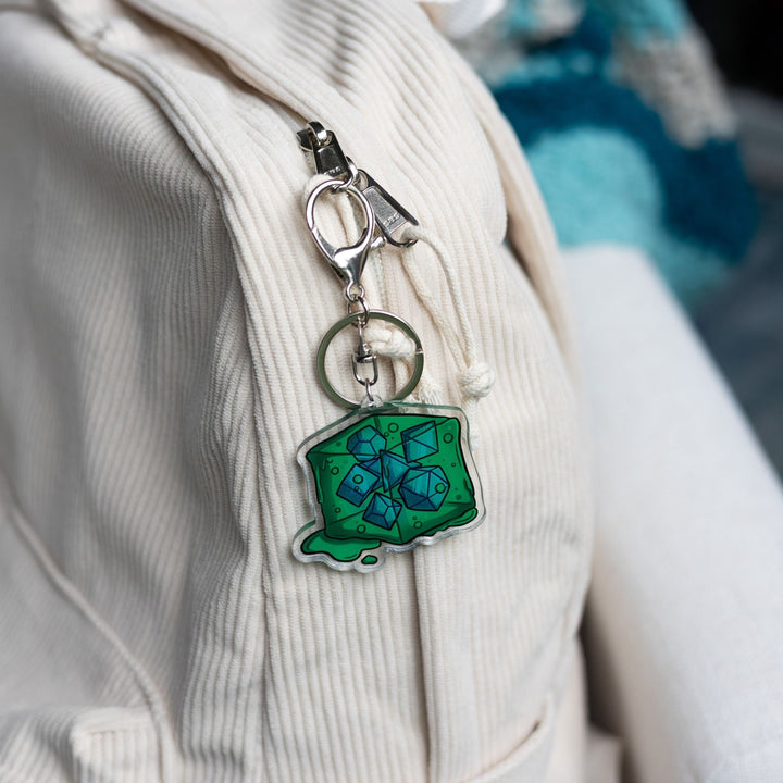 Acrylic keychain of a green gelatinous cube that is oozing and has DND dice inside of it clipped to a white backpack.
