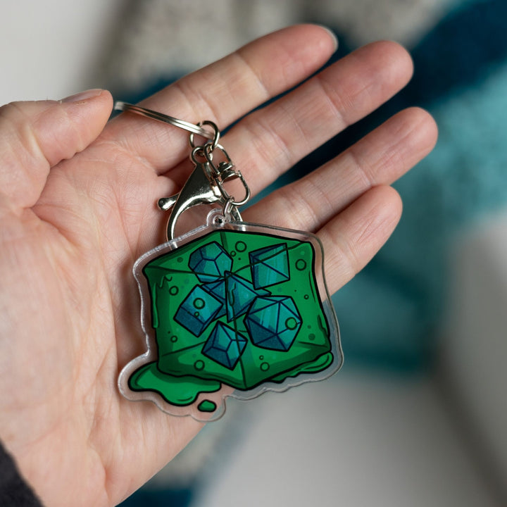  Acrylic keychain of a green gelatinous cube that is oozing and has DND dice inside of it held in a palm.