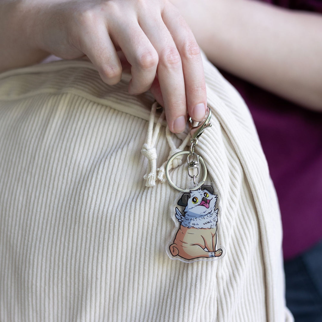 Acrylic keychain of Potroast, body and face of a pug with the beak and feathers of an owl  clipped to a white backpack. From Bookshops and Bonedust.