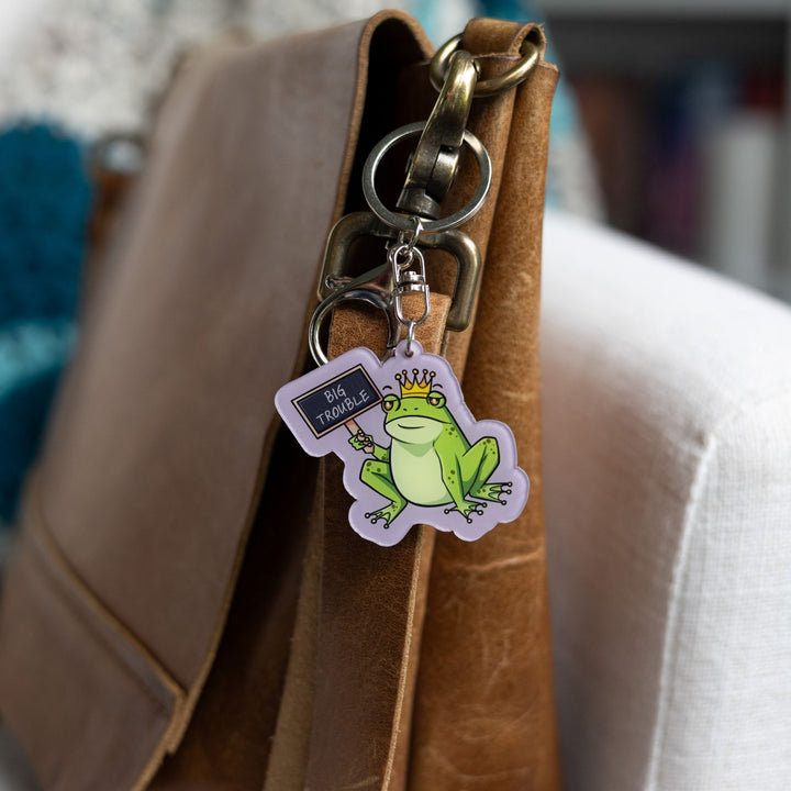 Acrylic keychain of a green frog wearing a crown holding up a chalk board sign that reads ‘Big Trouble’ clipped to a leather purse.
