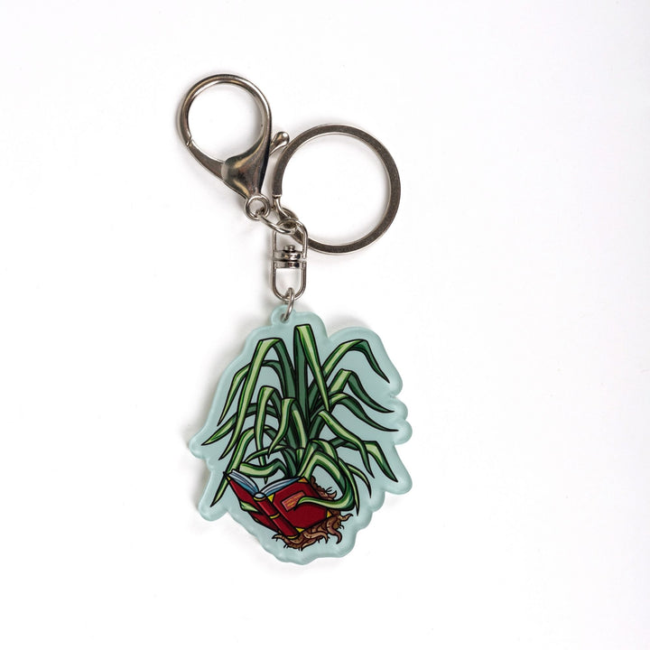 Acrylic keychain of a spider plant out of its pot holding a book with its leaves and reading it. From The Spellshop.