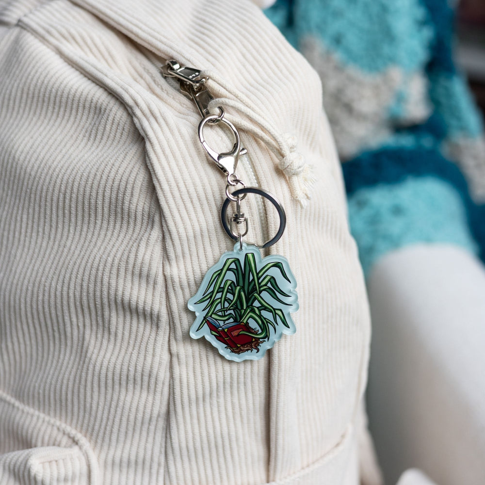 Acrylic keychain of a spider plant out of its pot holding a book with its leaves and reading it  clipped to a white backpack. From The Spellshop.
