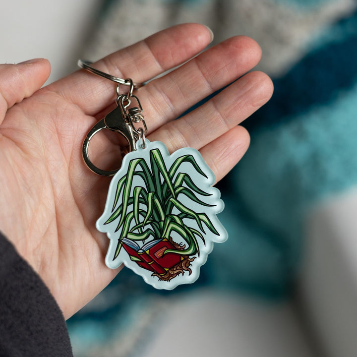 Acrylic keychain of a spider plant out of its pot holding a book with its leaves and reading it held in a palm. From The Spellshop.