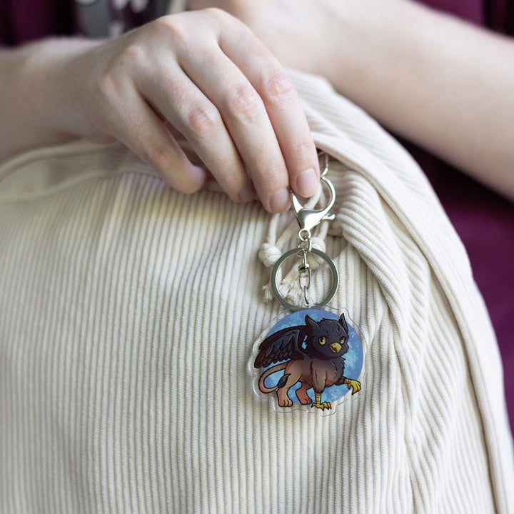 Acrylic Keychain of a baby griffon with a black head and wings and a brown body clipped to a white backpack. From Tomes & Tea.