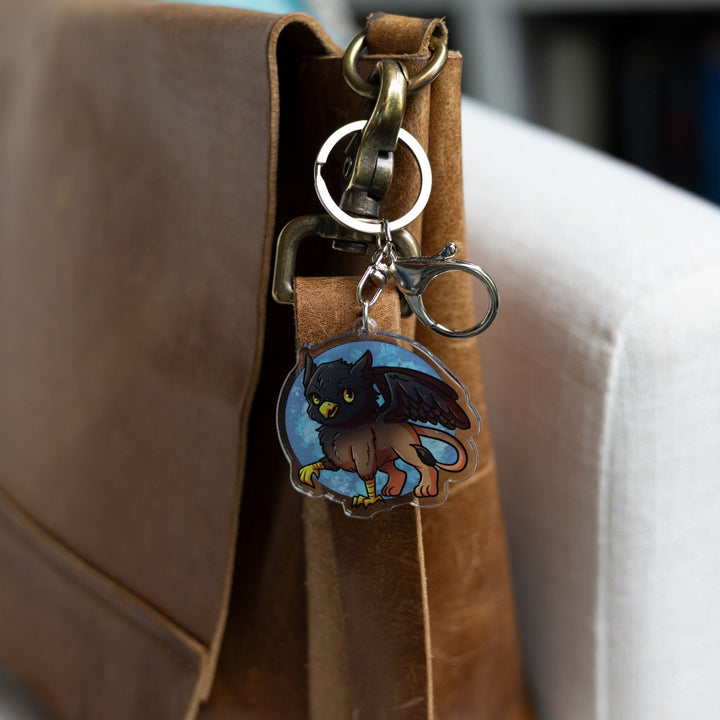 Acrylic Keychain of a baby griffon with a black head and wings and a brown body clipped to a leather purse. From Tomes & Tea.
