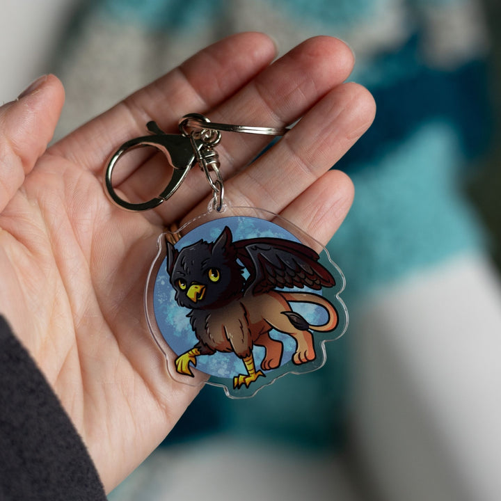 Acrylic Keychain of a baby griffon with a black head and wings and a brown body held in a palm. From Tomes & Tea