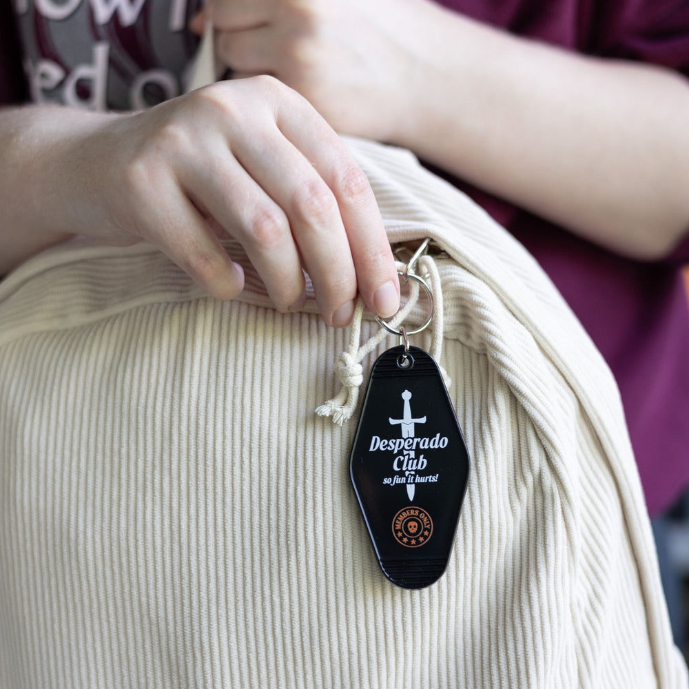 Black motel-style keychain with a silver dagger with the text ‘Desperado Club. So fun it hurts!’  clipped to a white backpack.
