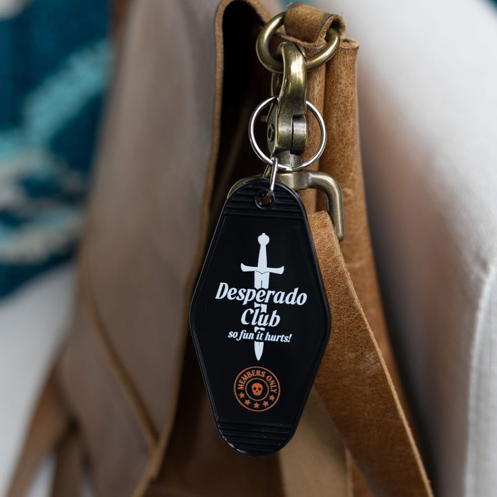 Black motel-style keychain with a silver dagger with the text ‘Desperado Club. So fun it hurts!’ clipped to a leather purse. 