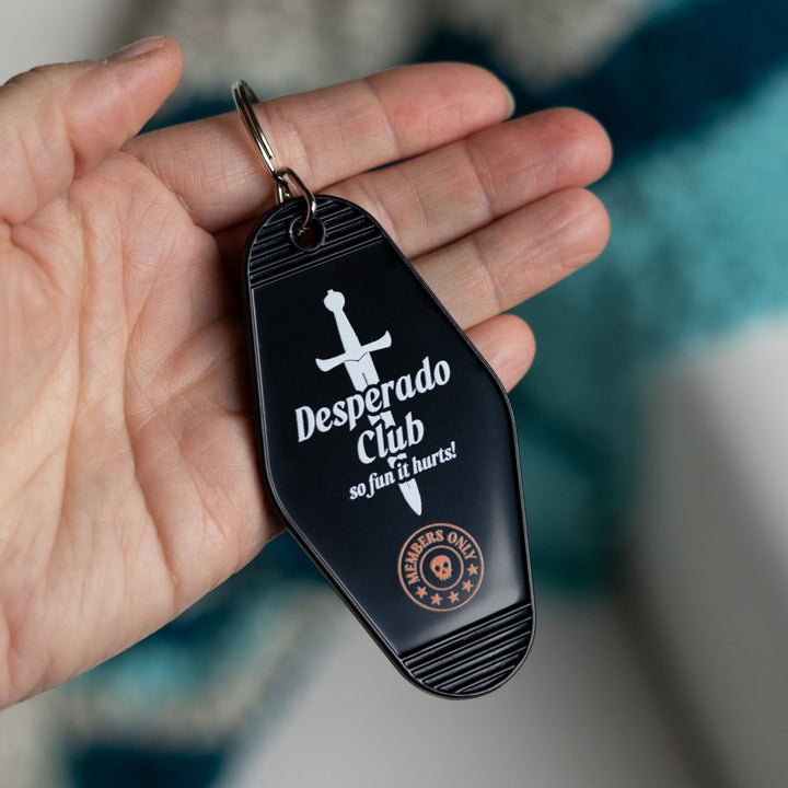 Black motel-style keychain with a silver dagger with the text ‘Desperado Club. So fun it hurts!’ held in a palm. From Dungeon Crawler Carl.
