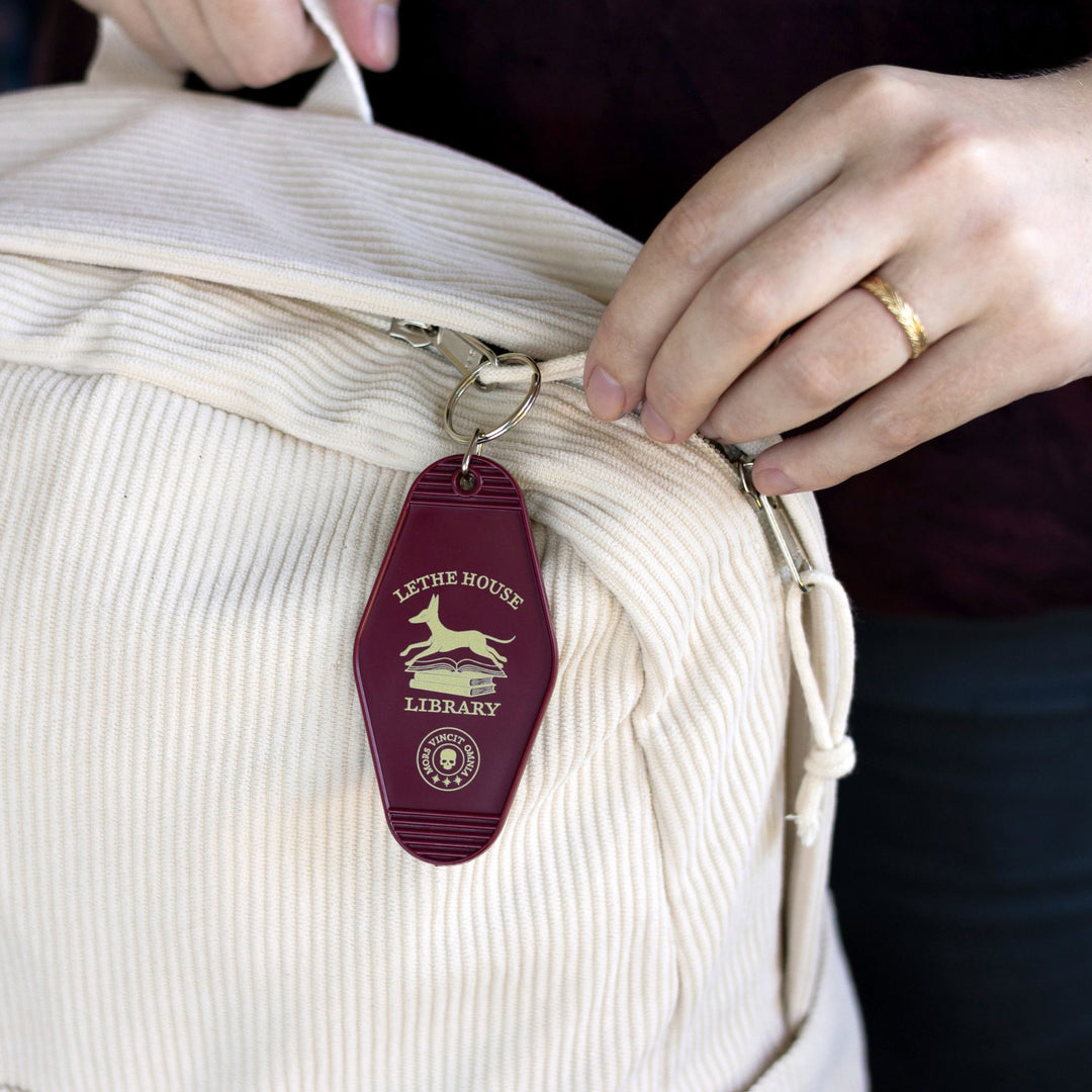Double sided motel keychain with an illustration of a jackal bounding over a stack of books that reads ‘Lethe House Library, Mors Irrumat Omnia.’ with dimensions displayed of 3 in by 1.75 in. On a beige back pack.