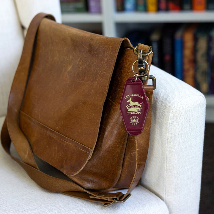 Double sided motel keychain with an illustration of a jackal bounding over a stack of books that reads ‘Lethe House Library, Mors Irrumat Omnia.’ On a leather bag.