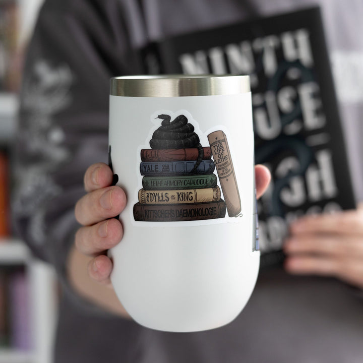 Sticker of stack of books from the Lethe House library from Ninth House by Leigh Bardugo with snake on top, on a white tumbler.
