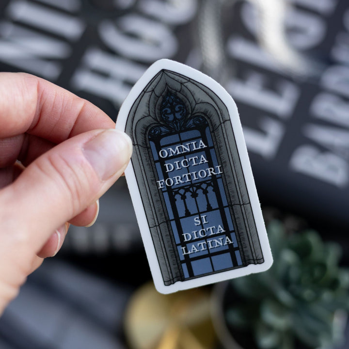Sticker of Sterling library window with text ‘Omnia Dicta Fortiori Si Dicta Latina’ held over Ninth House by Leigh Bardugo.
