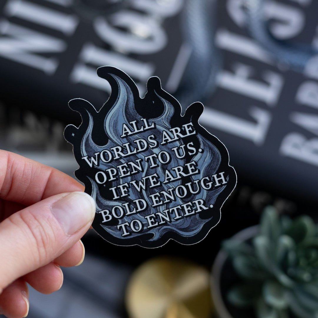  Quote sticker from Ninth House on blue flames that reads ‘ All worlds are open to us. If we are bold enough to enter,’ Held above Ninth House.