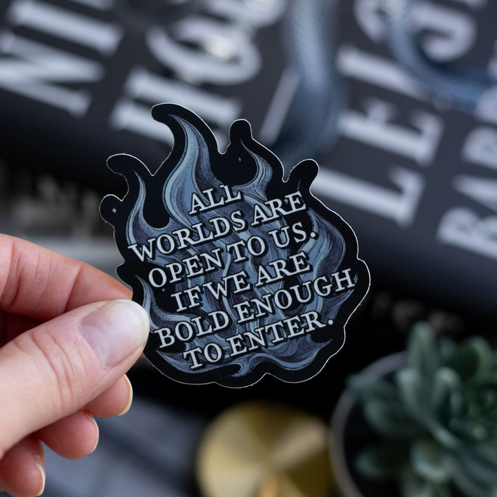  Quote sticker from Ninth House on blue flames that reads ‘ All worlds are open to us. If we are bold enough to enter,’ Held above Ninth House.
