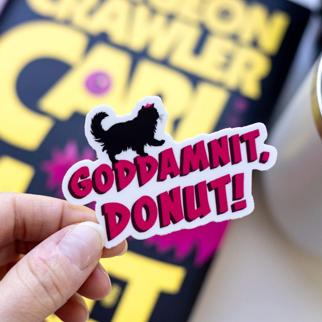 Sticker of 'Goddmanit, Donut!' in pink text with the silhouette of a prancing cat on top, held above Dungeon Crawler Carl.