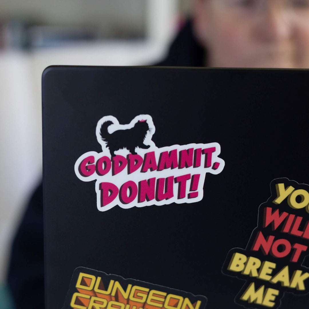 Sticker of 'Goddmanit, Donut!' in pink text with the silhouette of a prancing cat on top, on corner of a black laptop.