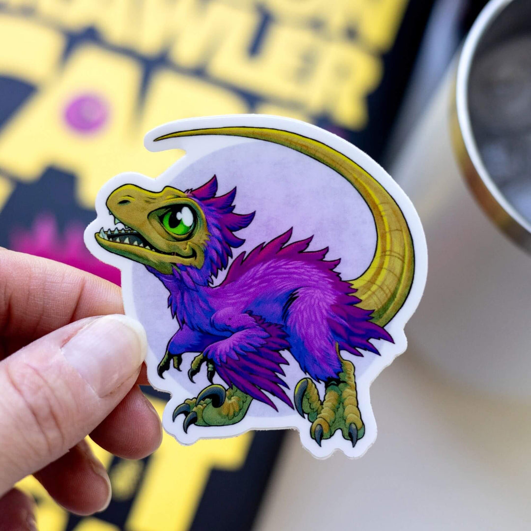  Sticker of Baby Mongo, a tiny, purple feathered velociraptor who has green eyes, and very sharp talons, held over Dungeon Crawler Carl.

