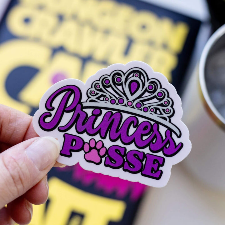 Sticker of ‘Princess Posse’ in purple letters with a tiara on top and a paw print where the o should be in Posse. Held over Dungeon Crawler Carl
