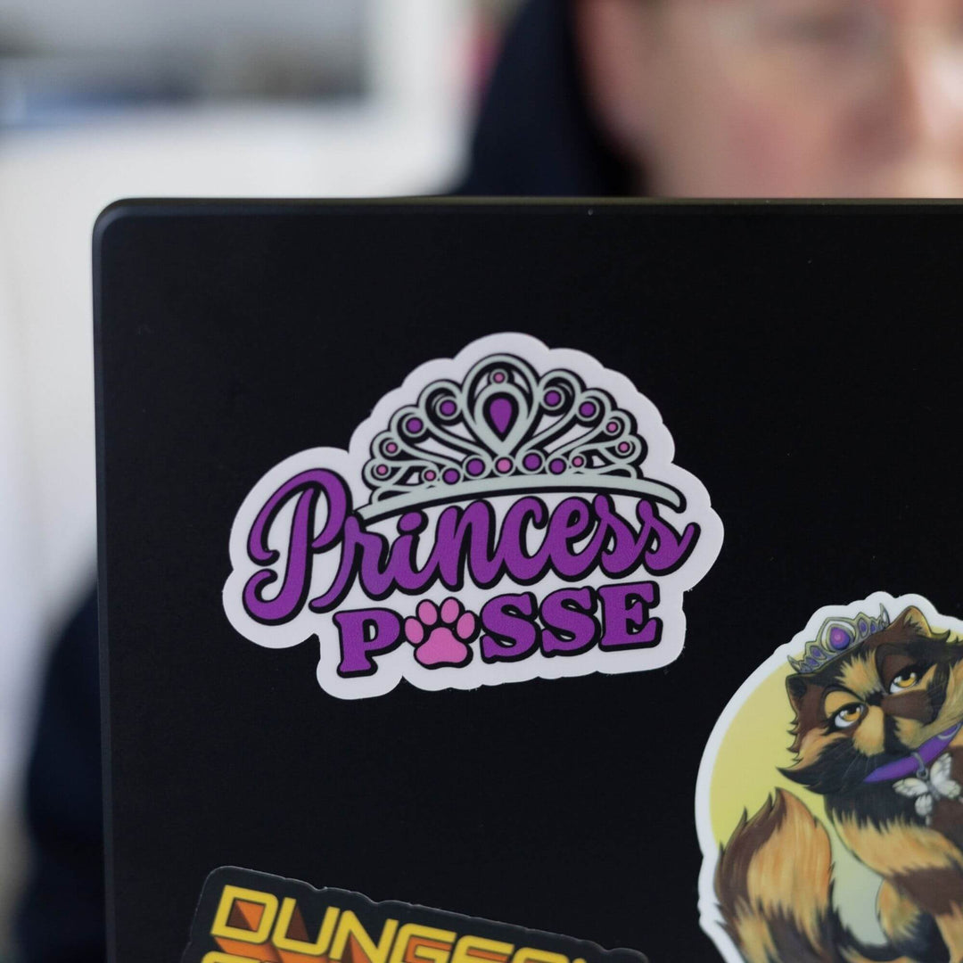 Sticker of ‘Princess Posse’ in purple letters with a tiara on top and a paw print where the o should be in Posse. On the corner of a black laptop.
