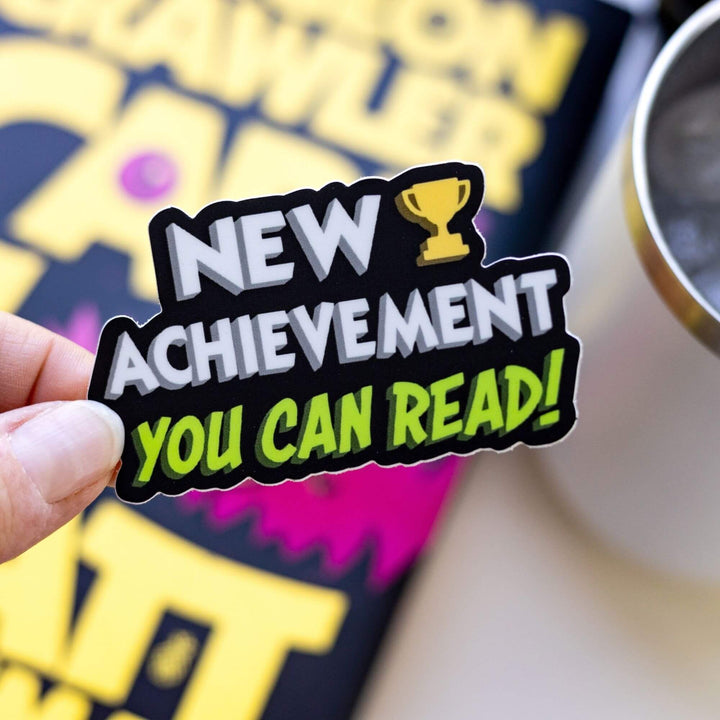  Sticker reads ‘New Achievement You Can Read!’ next to a trophy. Letters are white and green, outlined in black. Held over Dungeon Crawler Carl.
