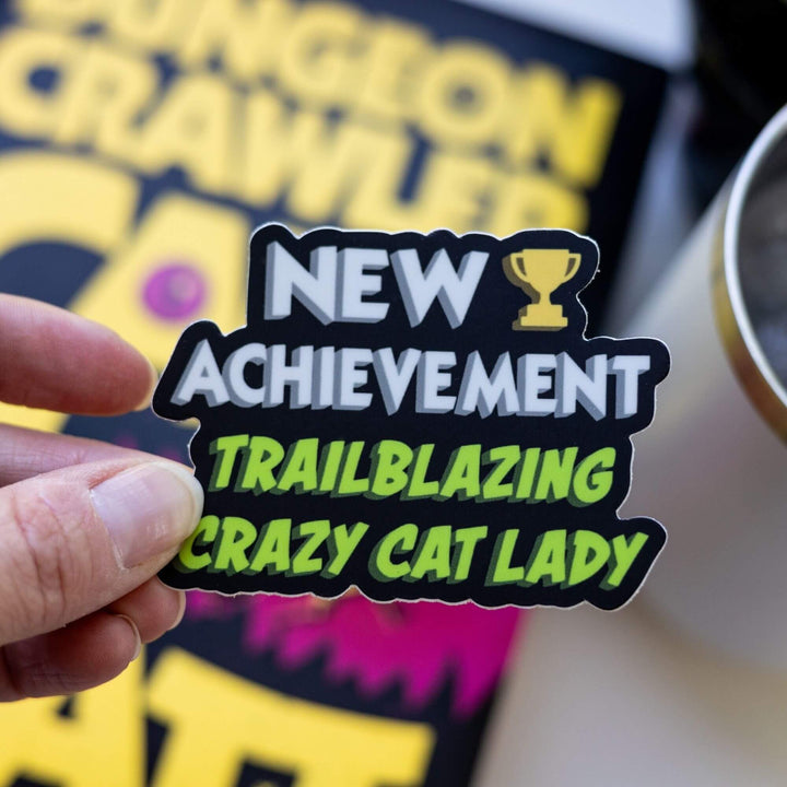 Sticker reads ‘New Achievement Trailblazing Crazy Cat Lady!’ next to a trophy. Letters are white and green, outlined in black. Held over Dungeon Crawler Carl.