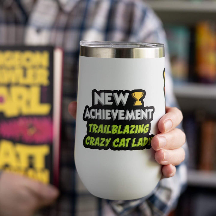 Sticker reads ‘New Achievement Trailblazing Crazy Cat Lady!’ next to a trophy. Letters are white and green, outlined in black. On a white tumbler.