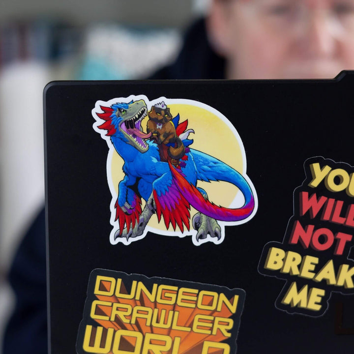 Sticker of Princess Donut in a saddle riding a fully grown Velociraptor, Mongo, who is roaring. On black laptop