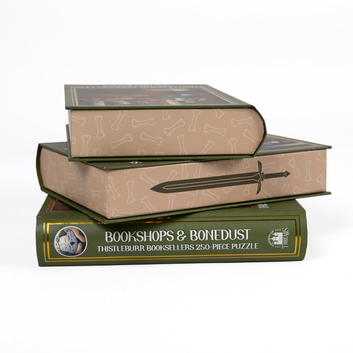 Stack of 3 puzzle boxes showing all sides. Beige edges with a sword and dog bones and the spine of the box reads Bookshops & Bonedust Thistleburr Booksellers 250-piece puzzle.