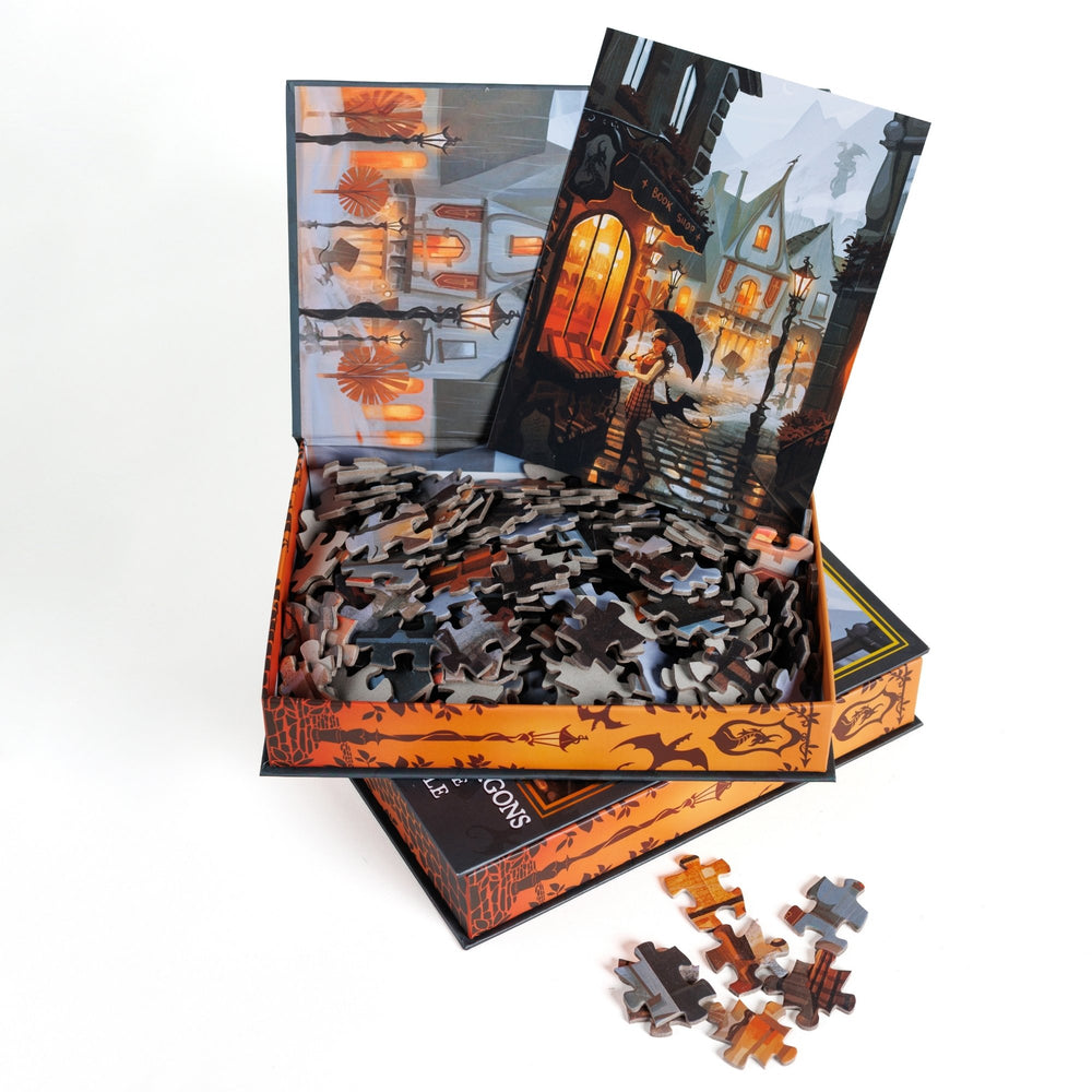 An open book-shaped box reveals puzzle pieces. Inner art on the top flap quaint buildings. An art print of a rainy day street scene outside a bookstore atop the pieces.