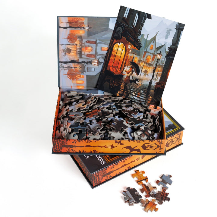 An open book-shaped box reveals puzzle pieces. Inner art on the top flap quaint buildings. An art print of a rainy day street scene outside a bookstore atop the pieces.