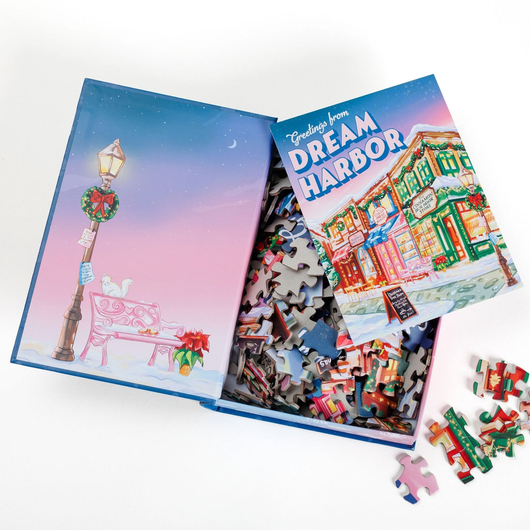 An open book-shaped box reveals puzzle pieces. Inner art on the top flap lamppost and bench. An art print of Main Street decorated for the holidays atop the pieces.