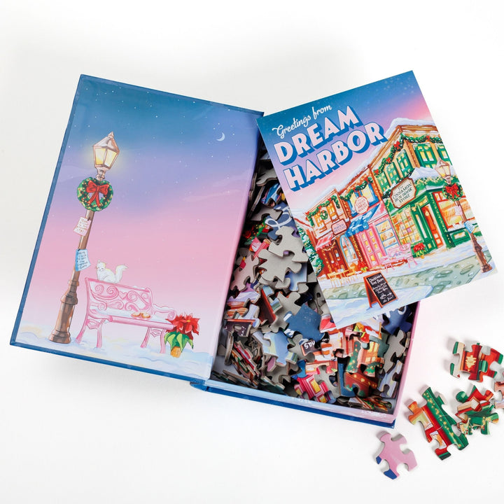 An open book-shaped box reveals puzzle pieces. Inner art on the top flap lamppost and bench. An art print of Main Street decorated for the holidays atop the pieces.
