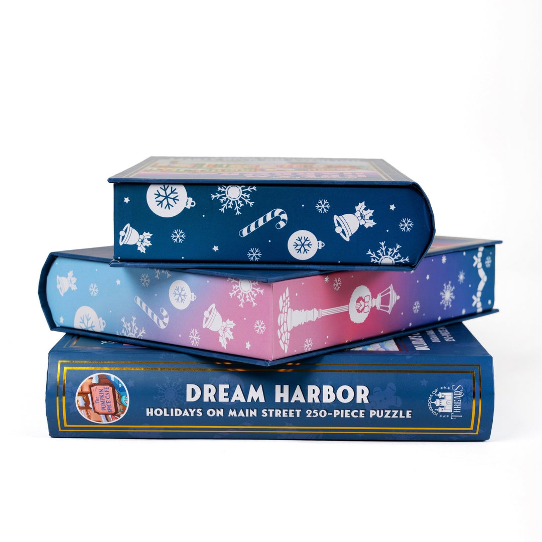 Stack of 3 puzzle boxes showing all sides. Pink and blue edges with wintery designs and the spine of the box reads Dream Harbor Holidays on Main Street 250-piece puzzle.