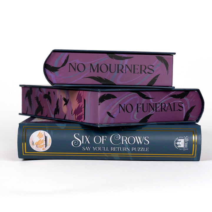 Stack of 3 puzzle boxes showing all sides. Purple edges with crows and feathers. On different sides ‘No mourners’ ‘No funerals.’ The spine of the box reads Six of Crows Say You’re return puzzle.