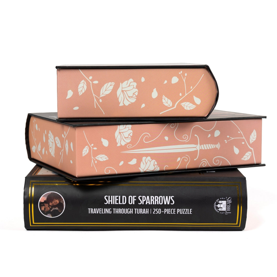 Stack of 3 puzzle boxes showing all sides. Peach edges with white roses and daggers and the spine of the box reads ‘Shield of Sparrows Traveling Through Turah  250-piece puzzle.’