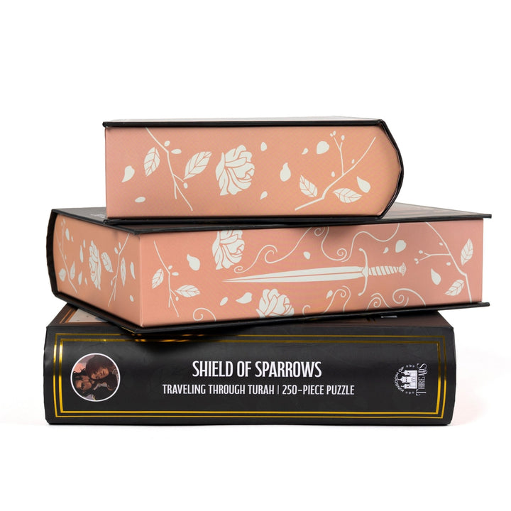 Stack of 3 puzzle boxes showing all sides. Peach edges with white roses and daggers and the spine of the box reads ‘Shield of Sparrows Traveling Through Turah  250-piece puzzle.’