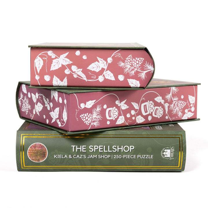 Stack of 3 puzzle boxes showing all sides. Pink edges with white raspberries and jam jars. The spine of the box reads The Spellshop Kiela and Caz’s Jam Shop 250-piece puzzle.