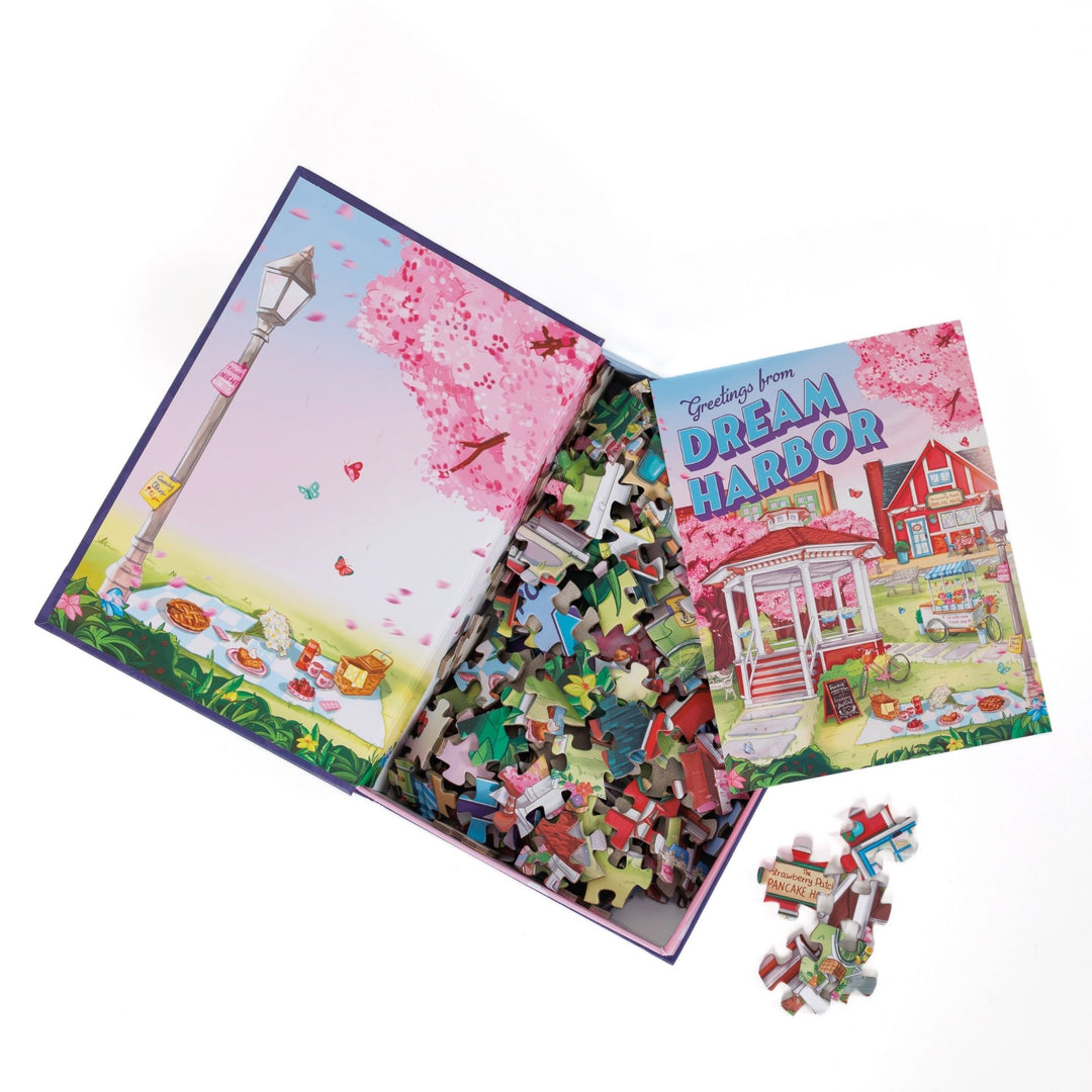 n open book-shaped box reveals puzzle pieces. Inner art on the top flap of a picnic under cherry blossoms. An art print of a gazebo in spring atop the pieces.