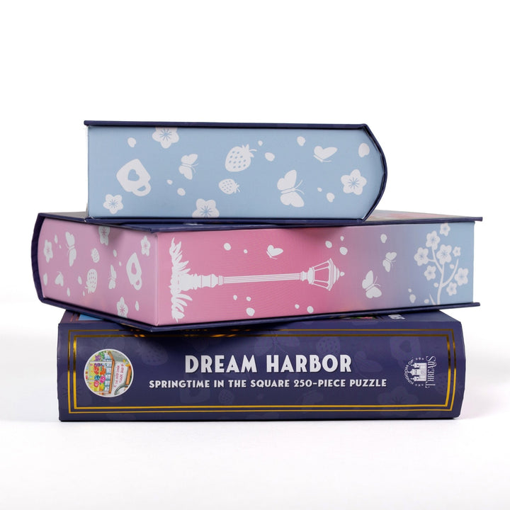 Stack of 3 puzzle boxes showing all sides. Pastel blue and pink edges with white butterflies. The spine of the box reads  Dream Harbor Springtime in the Square 250-piece puzzle.