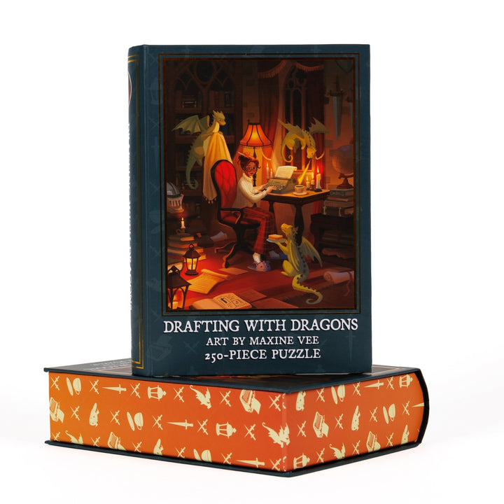 Book-shaped dark blue box for ‘Drafting with Dragons Art by Maxine Vee 250-piece puzzle.’ Art on the front is of a woman at a writing desk with three small dragons.