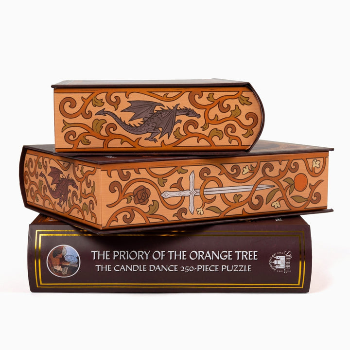 Stack of three puzzle boxes showing all sides. Orange edges with a dragon, ornate orange vines. The spine of the box reads ‘The Priory of the Orange Tree The Candle Dance 250-piece puzzle.’