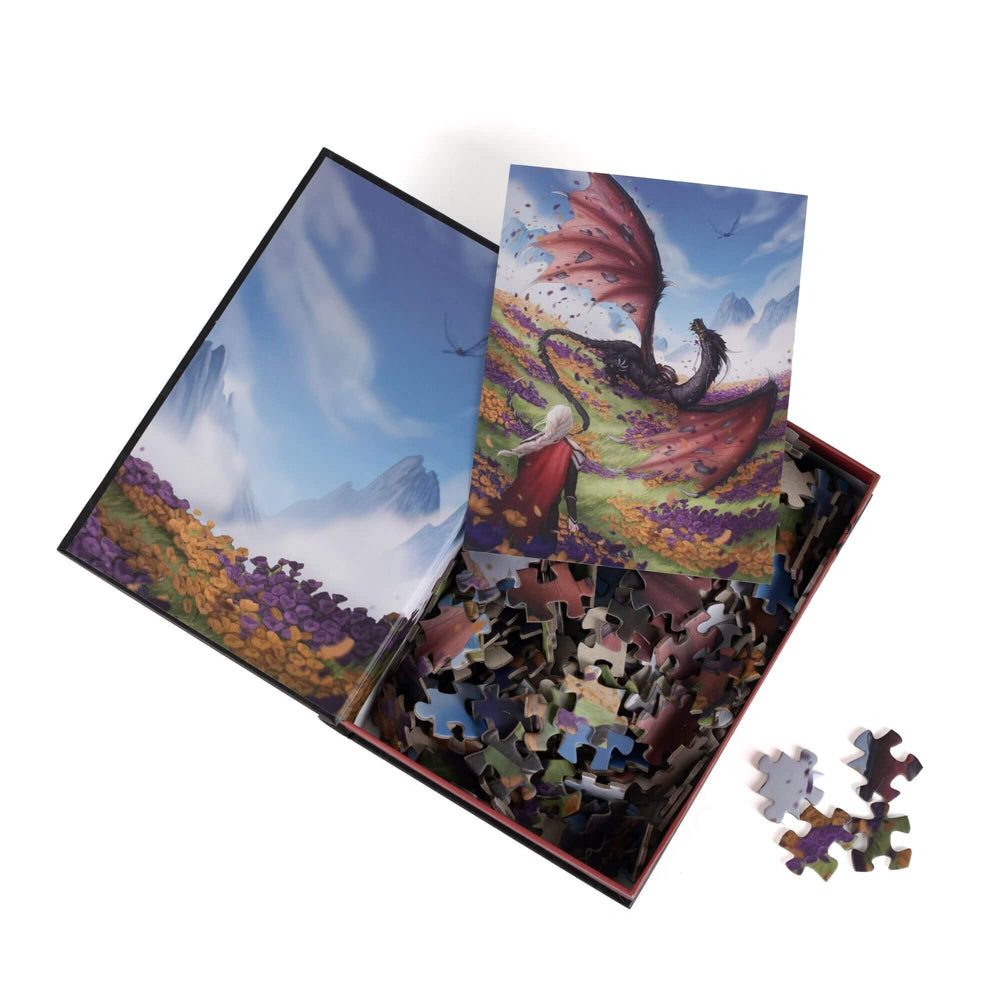 An open book-shaped box reveals puzzle pieces. Inner art on the top flap of a mountain scape. An art print of Manon & Abraxos atop the pieces.