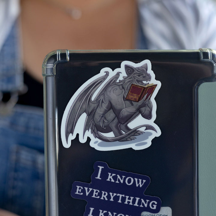 Sticker of a gargoyle reading a red, upside down book on a black laptop. Character is Bartholomew from The Knight and the Moth.