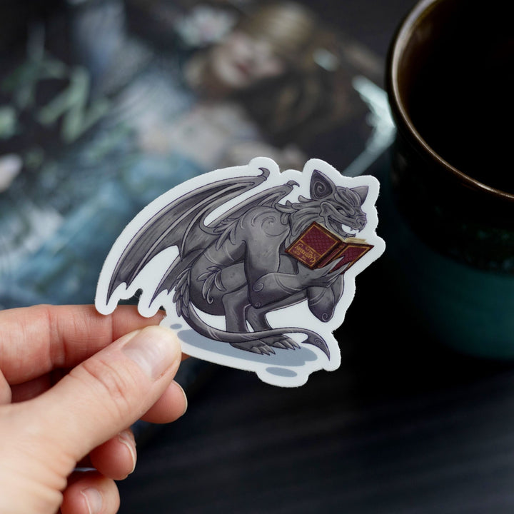 Sticker of a gargoyle reading a red, upside down book held over The Knight and the Moth by Rachel Gillig. The character is Bartholomew.