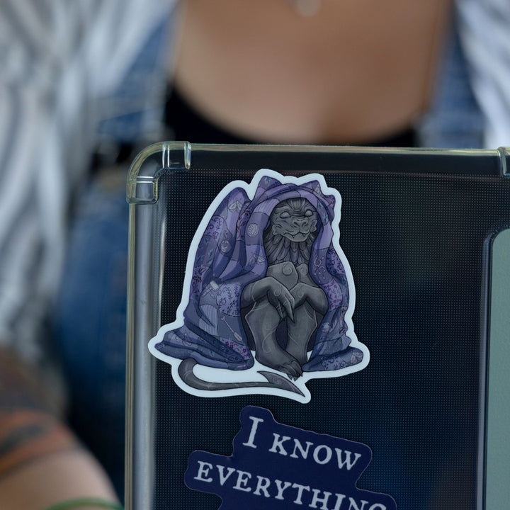 Sticker of a gargoyle sitting underneath a purple blanket on a black laptop. Character is Bartholomew from The Knight and the Moth.