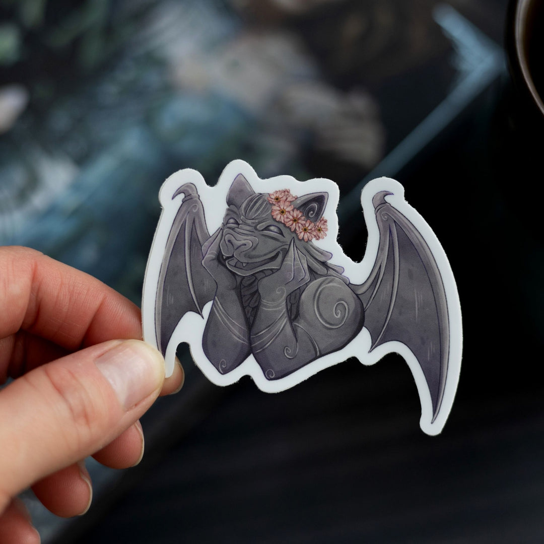 Sticker of a smiling gargoyle wearing a flower crown held over The Knight and the Moth by Rachel Gillig. The character is Bartholomew.